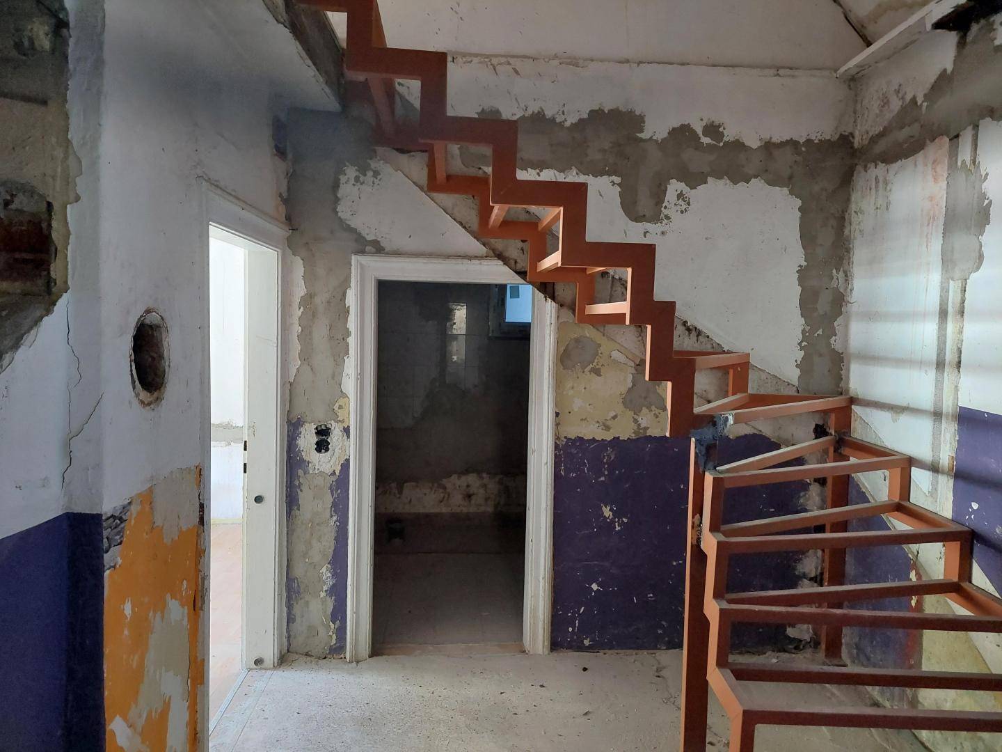 Innenraum im Rohbau mit freiliegenden Wänden, Türöffnungen und einer modernen orangefarbenen Metalltreppe.