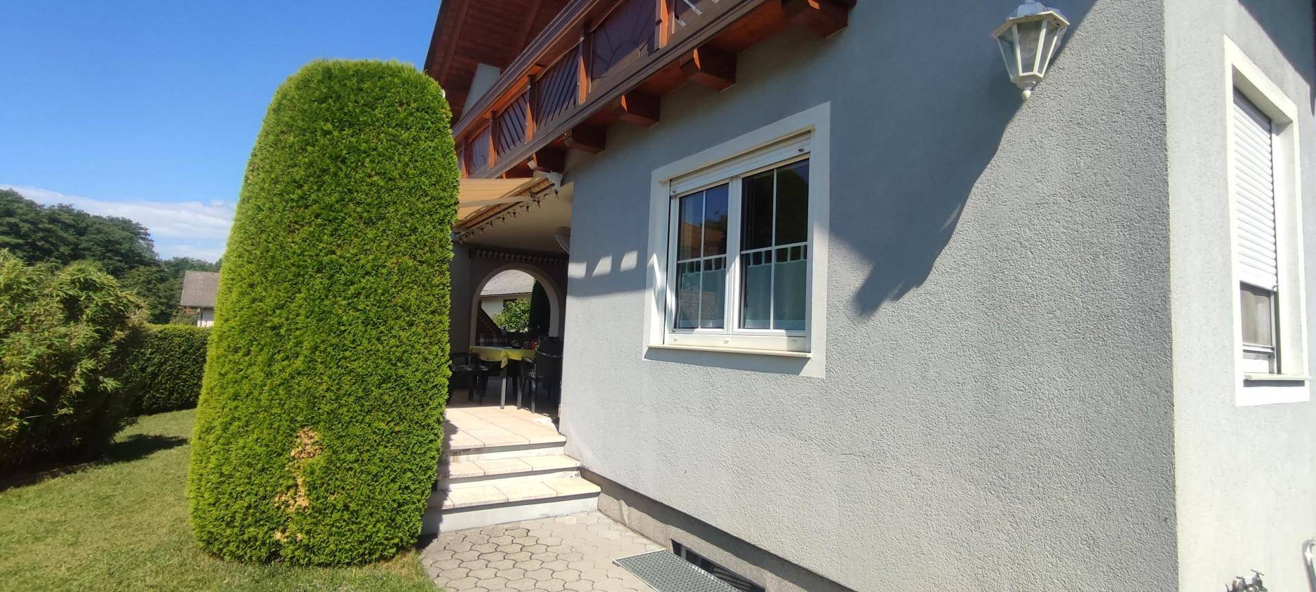 Hausfassade mit Gartenbereich, gepflegter Hecke und einem überdachten Sitzplatz im Freien.
