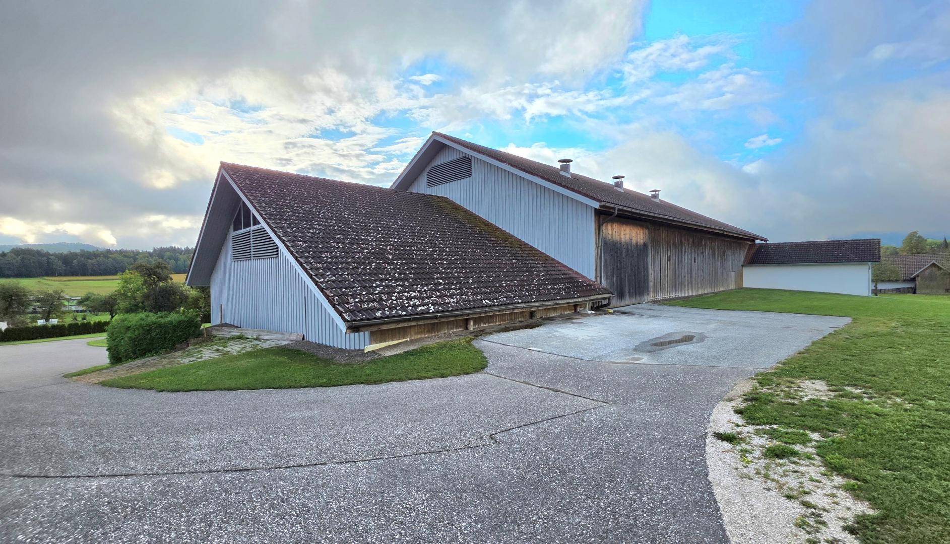 Seitenansicht eines großen Scheunengebäudes mit Holzwänden und einem weitläufigen Vorplatz.