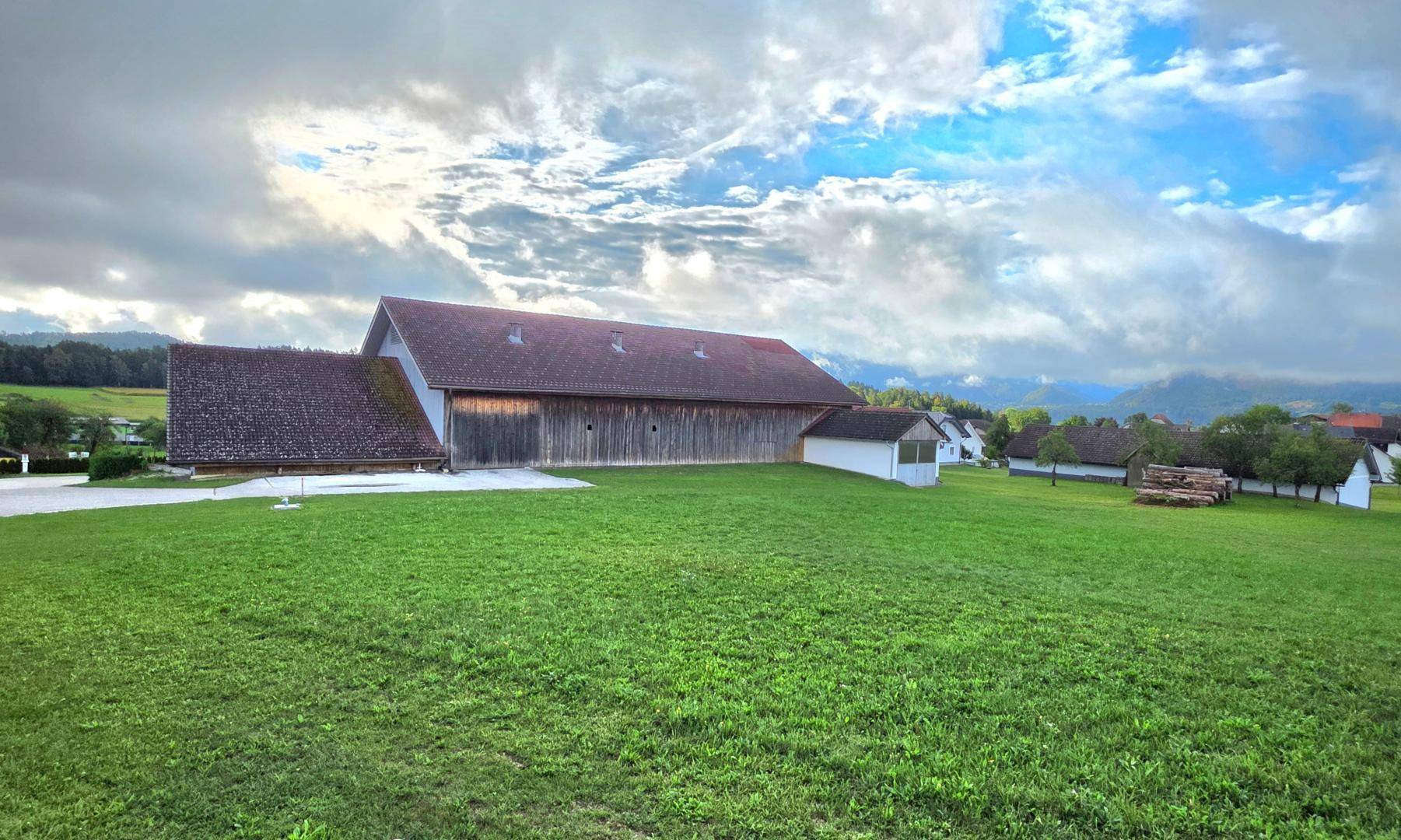 Großes Scheunengebäude umgeben von weiten grünen Feldern und ländlicher Landschaft unter bewölktem Himmel.