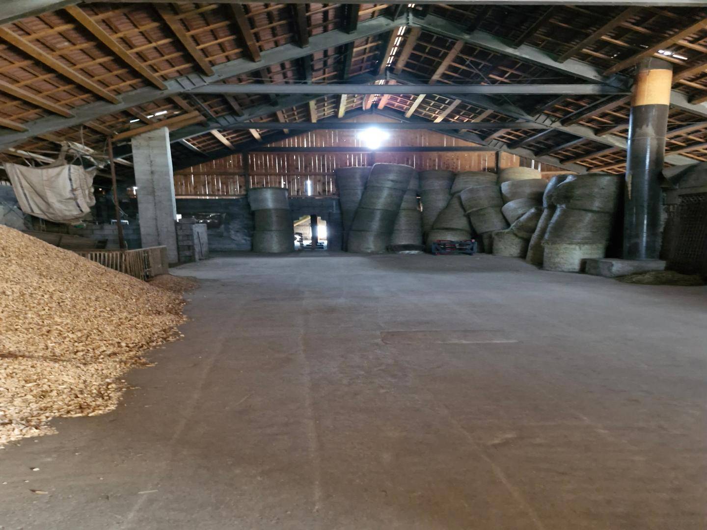Großer Lagerraum mit Holzbalken, Betonboden und gelagerten Heuballen sowie Getreidehaufen.