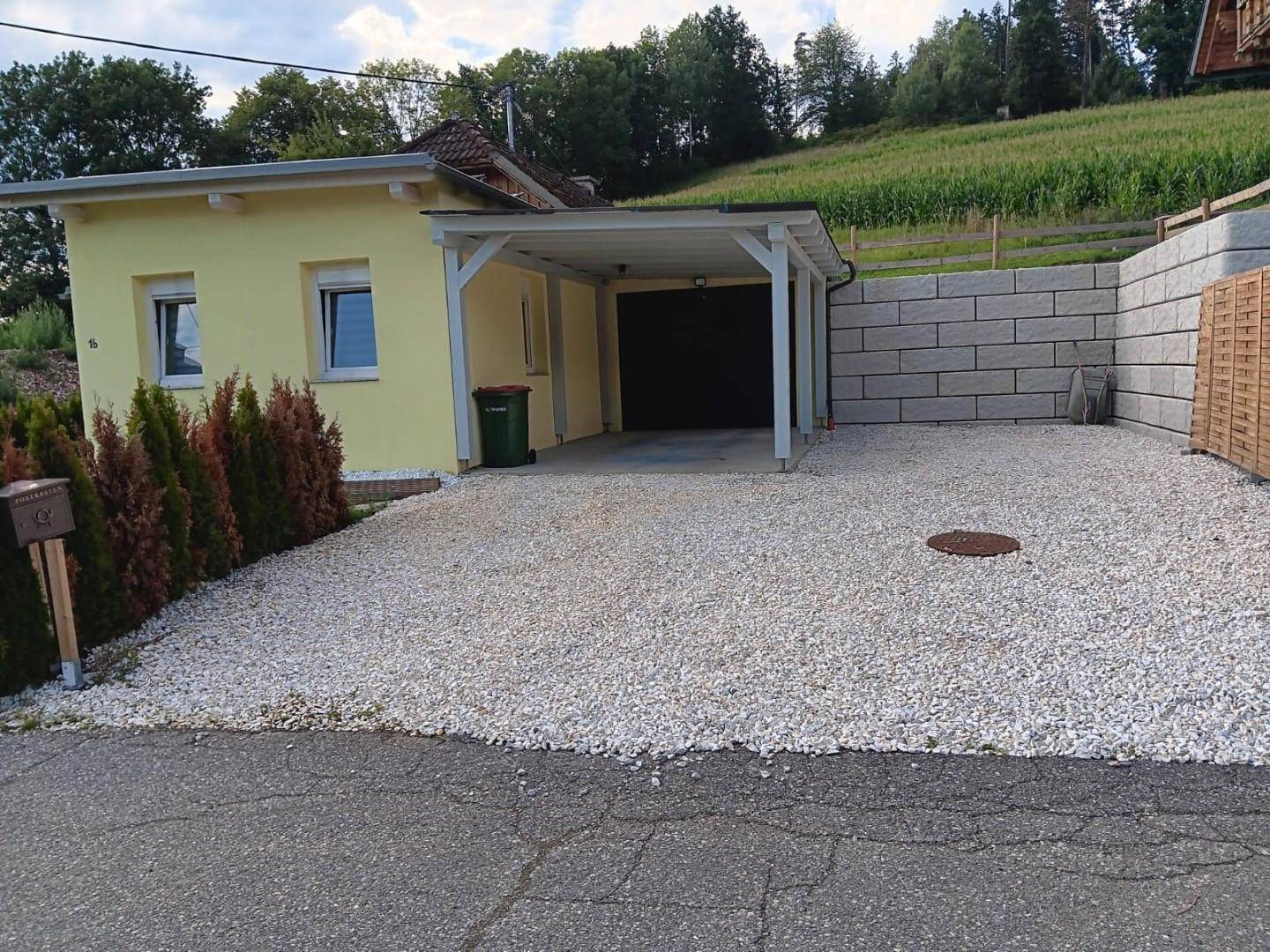 Einfamilienhaus mit Schotterauffahrt, Carport und einer Stützmauer im Hintergrund.