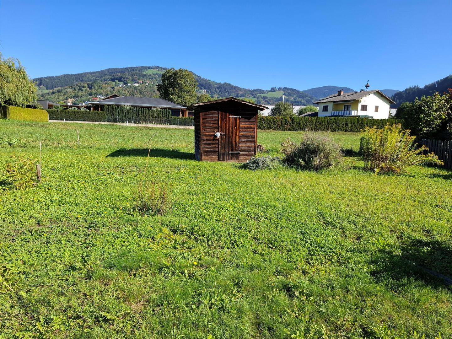 Weitläufige Rasenfläche mit einem kleinen Holzschuppen und Blick auf die umliegende Natur und Häuser.
