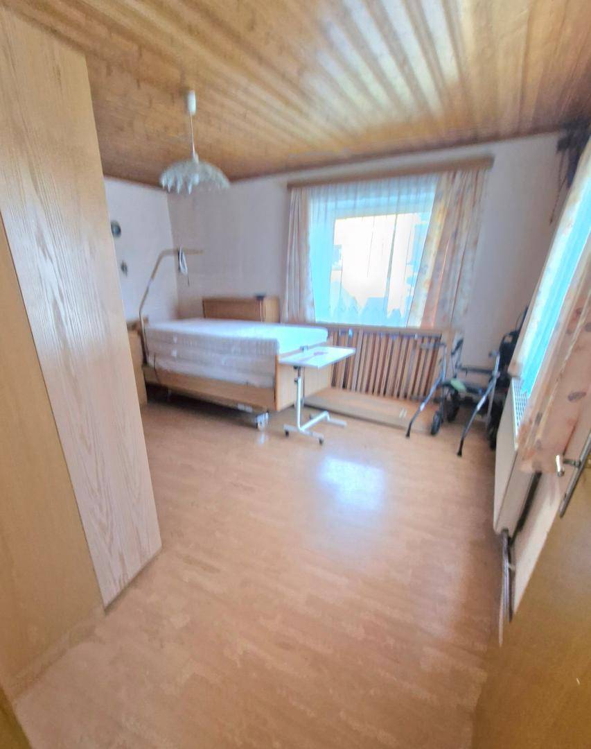 Schlafzimmer mit Pflegebett, Holzdecke und Laminatboden, hell durch Fenster.