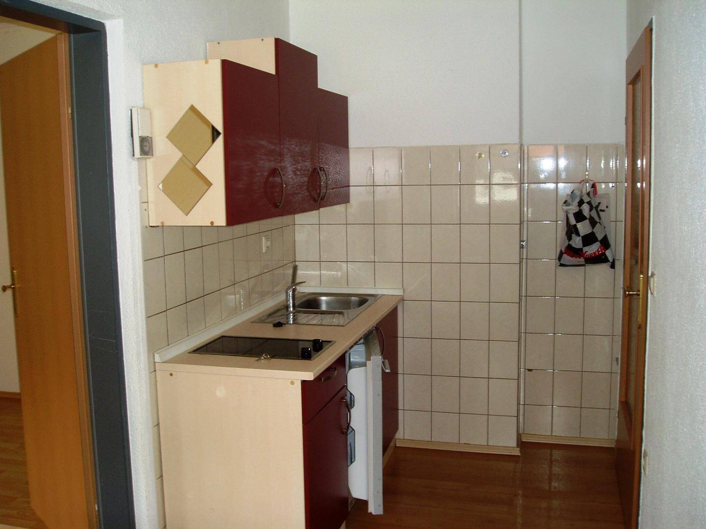 Kleine Küchenzeile mit Spüle, Kochfeld und Hängeschränken in Rot und Beige.
