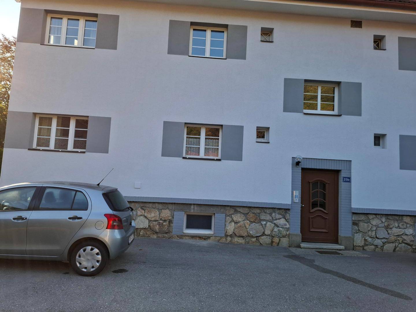 Fassade eines Mehrfamilienhauses mit Parkplatz, Steinsockel und grauen Akzenten.