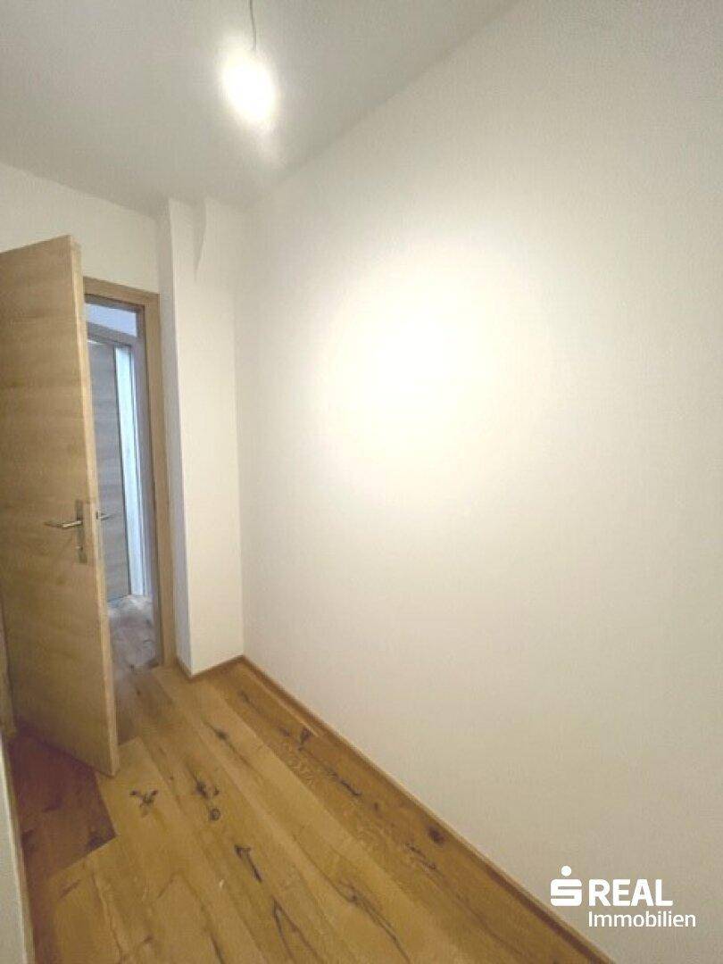 Schmaler Flur mit Holzboden und einer offenen Tür, die zu einem weiteren Raum führt.