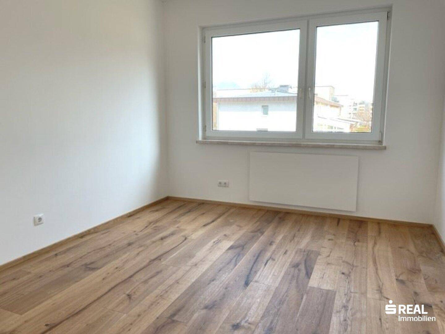 Heller Raum mit großem Fenster, das viel Tageslicht hereinlässt, und einem warmen Holzfußboden.