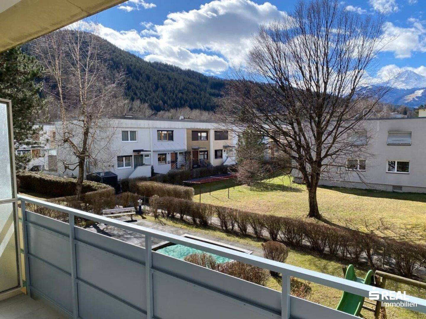 Balkonblick auf einen grünen Innenhof mit Spielplatz und die beeindruckende Bergkulisse.