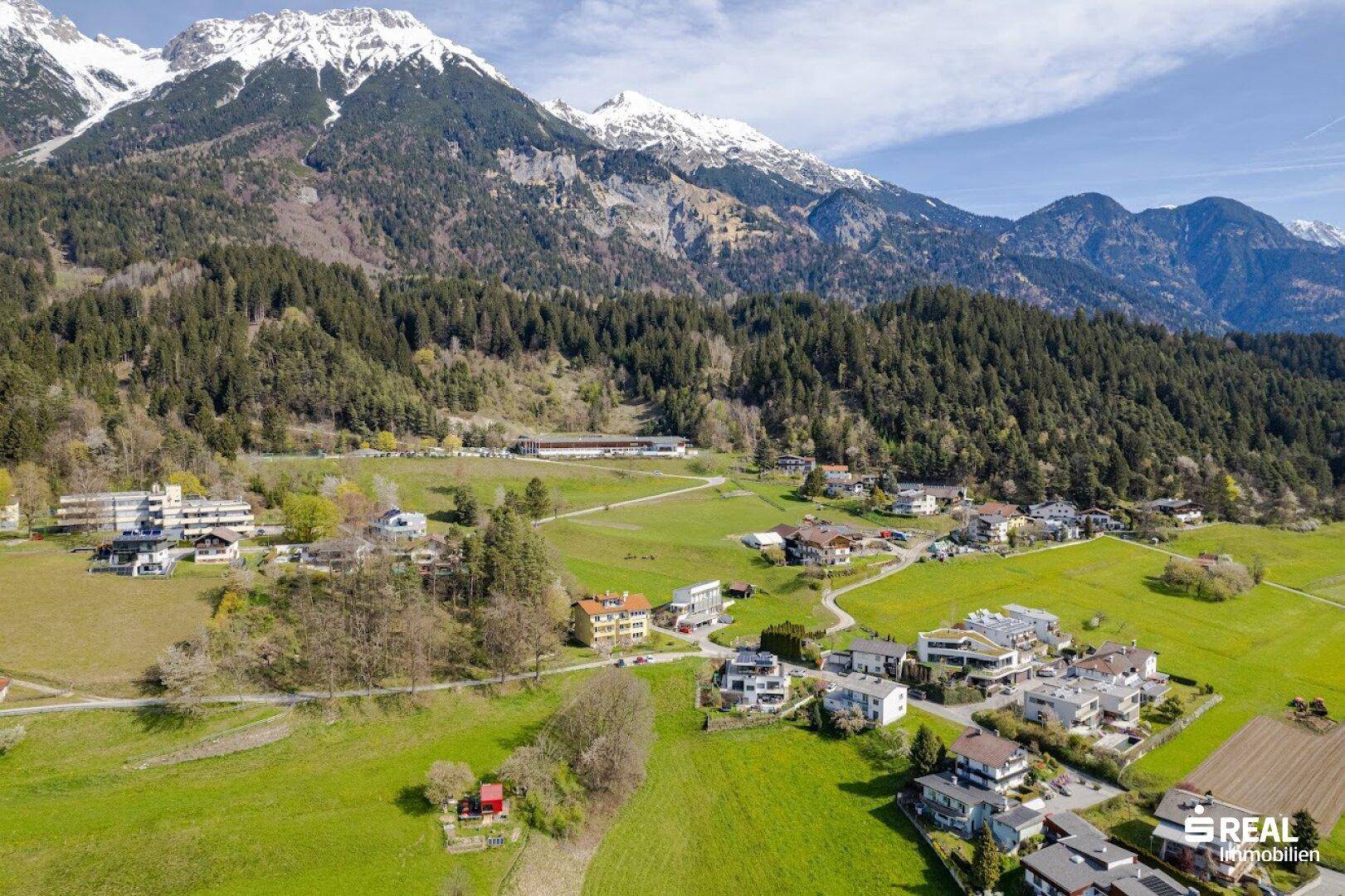 Luftaufnahme eines Wohngebiets am Fuße bewaldeter Berge mit grünen Wiesen und Häusern.