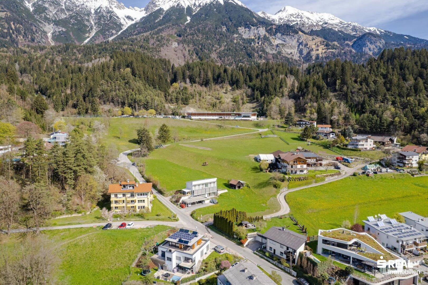Weitläufige Luftaufnahme einer Wohnsiedlung mit grünen Feldern und bewaldeten Bergen im Hintergrund.