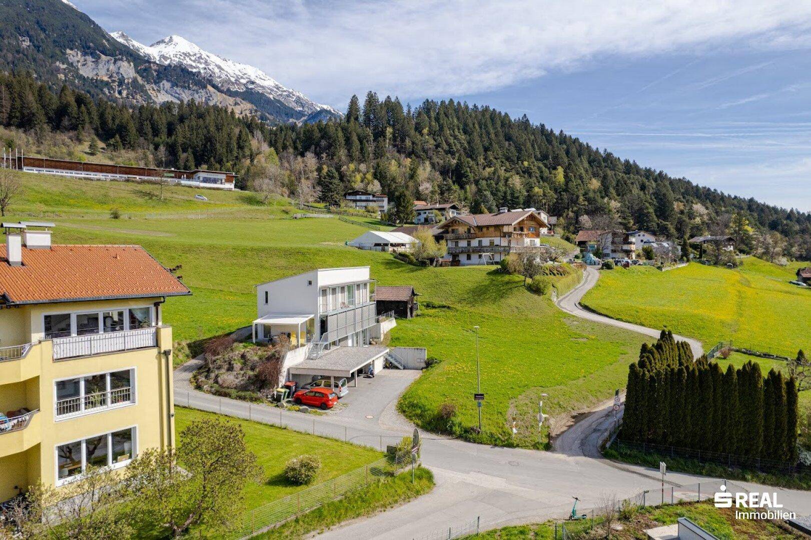 Blick auf eine Wohnsiedlung am Hang mit modernen Gebäuden und bewaldeten Bergen im Hintergrund.