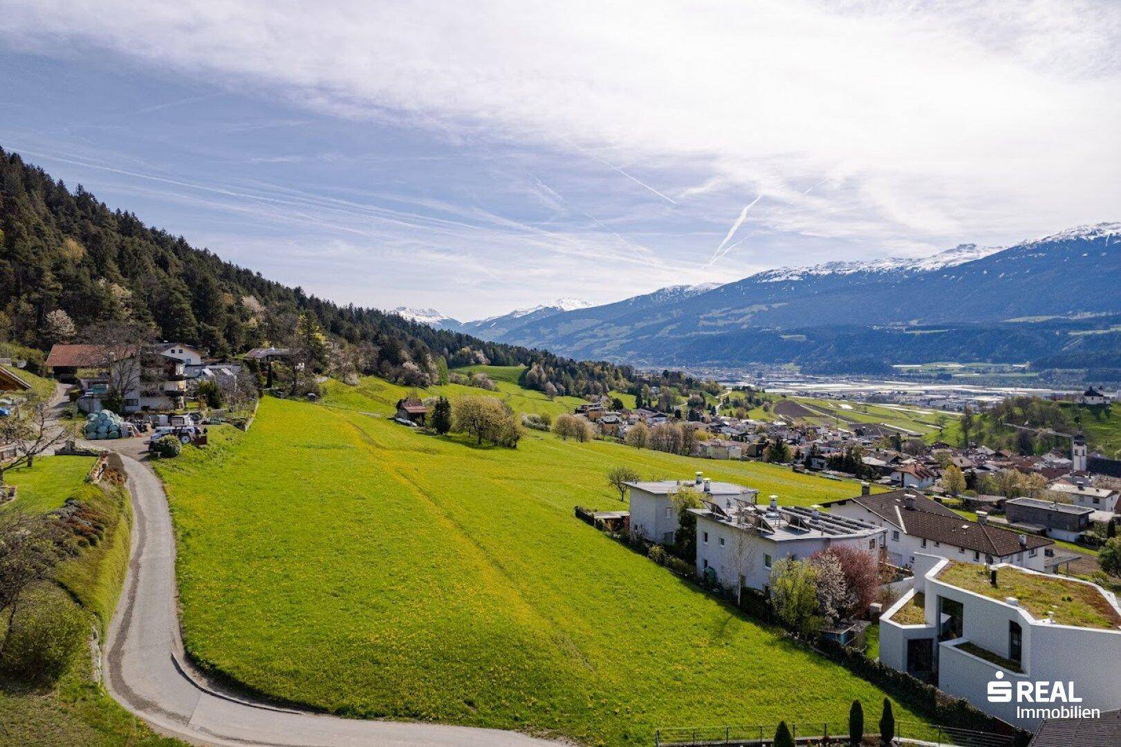 Weitläufiger Blick über grüne Hügel, eine Wohnsiedlung und die umliegende Berglandschaft.