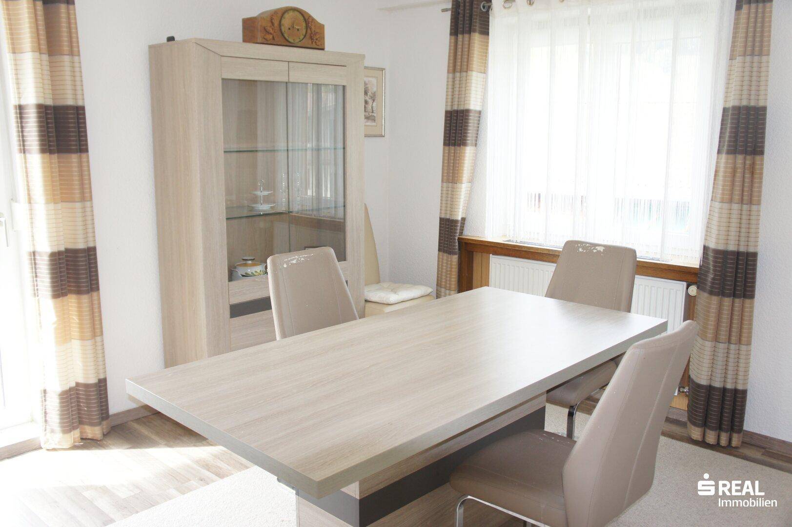 Esszimmer mit hellem Holztisch, Vitrinenschrank und Blick durch das Fenster.