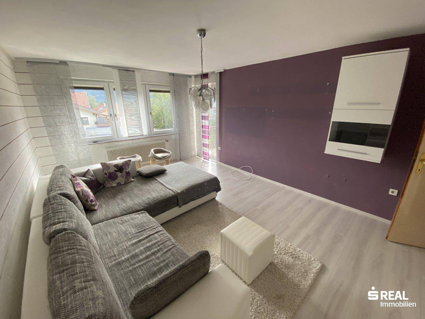 Geräumiges Wohnzimmer mit großer Couch, lila Akzentwand und hellem Laminatboden.