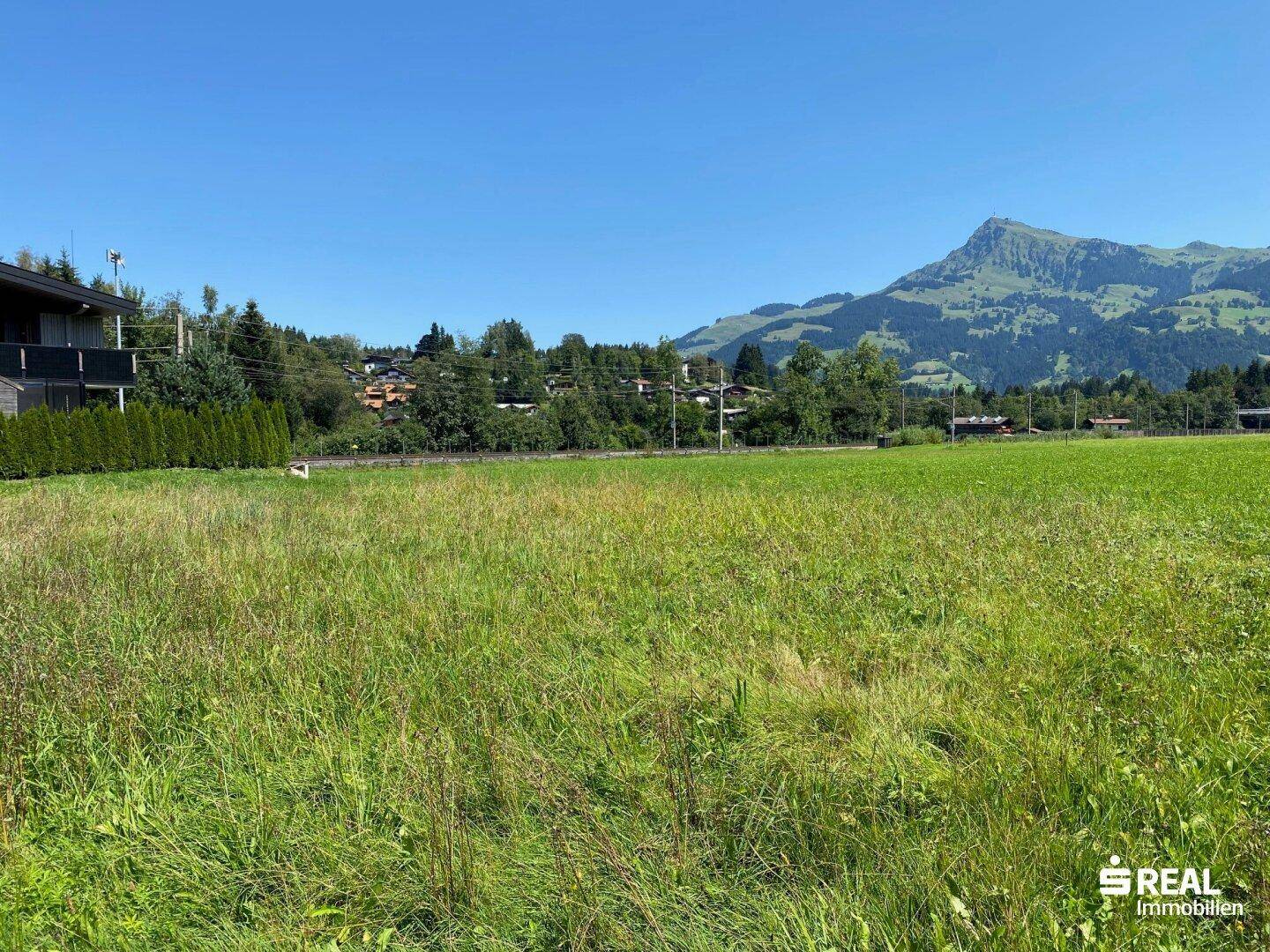 Weite grüne Fläche mit einem Wohnhaus, einer Hecke und einer beeindruckenden Bergkulisse im Hintergrund.