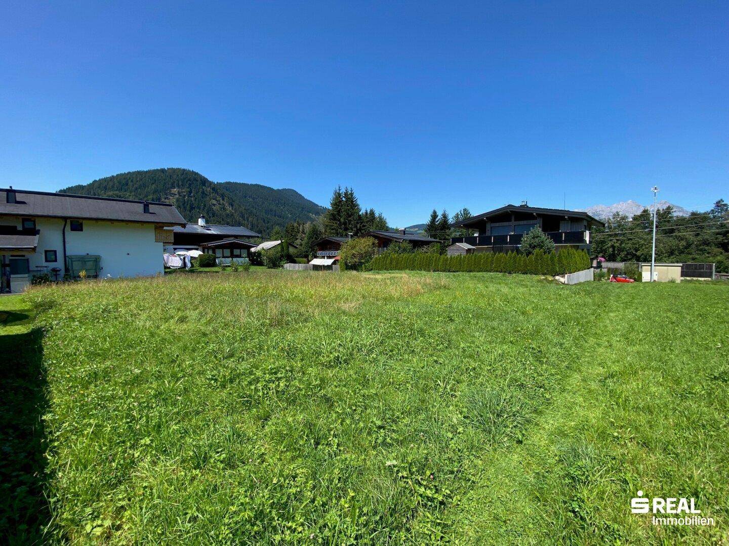 Grüne Wiese mit umliegenden Wohnhäusern und bewaldeten Hügeln im Hintergrund bei klarem Wetter.