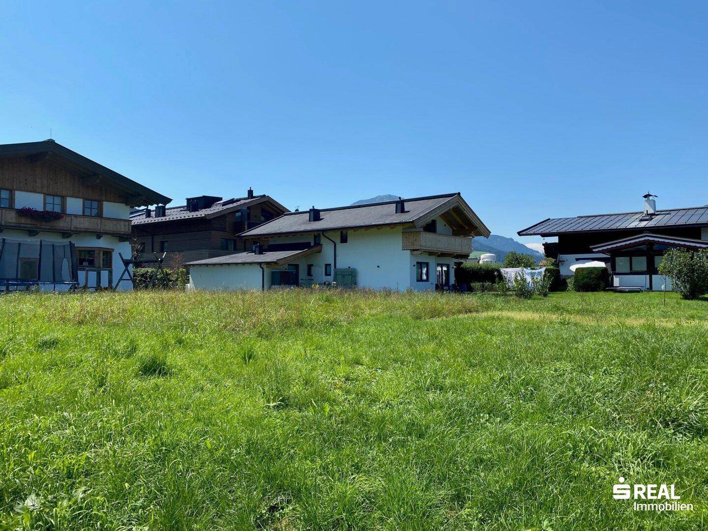 Offene Grünfläche mit traditionellen Wohnhäusern und Bergsilhouette unter einem wolkenlosen Himmel.