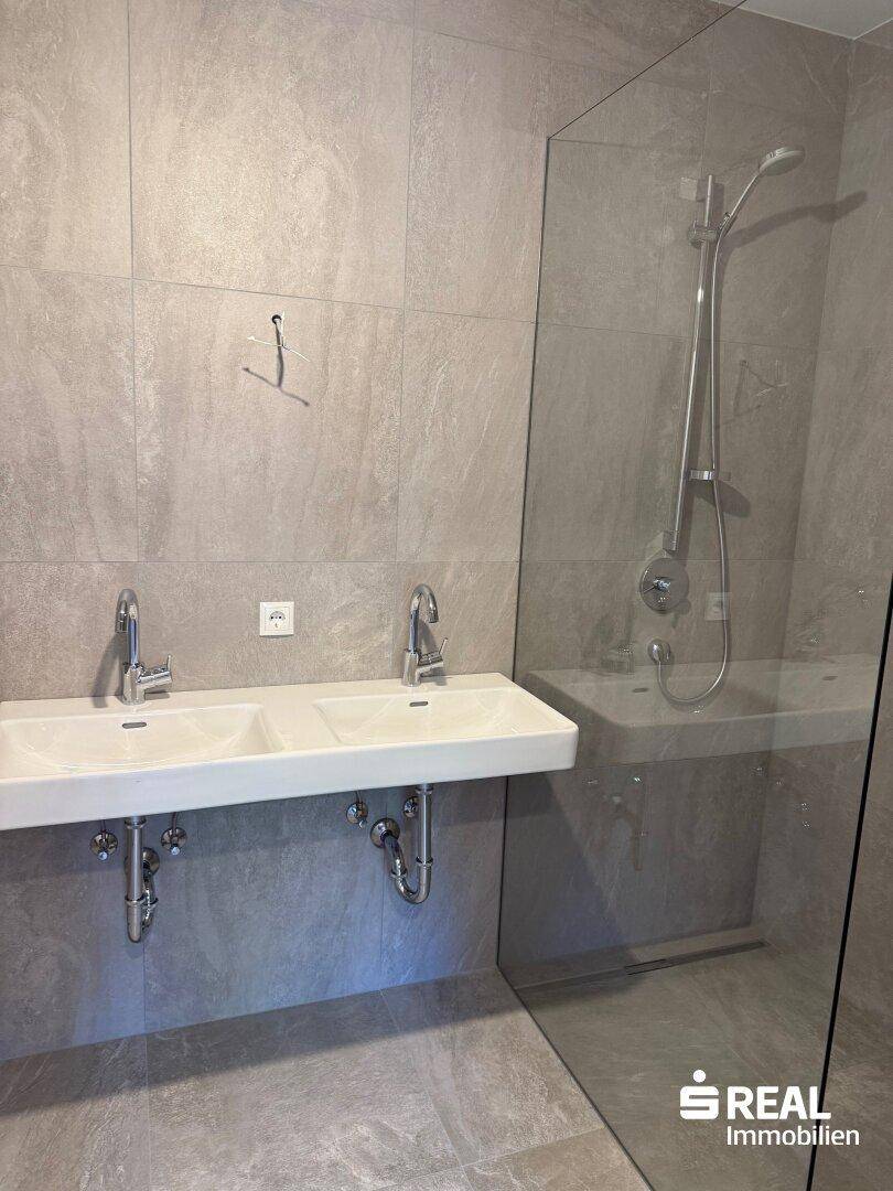 Modernes Badezimmer mit grauen Fliesen, Doppelwaschbecken und einer begehbaren Dusche.