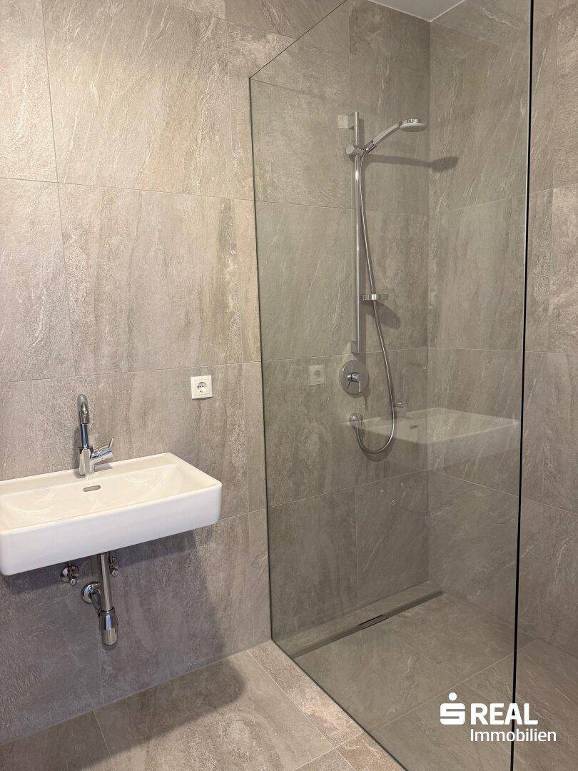 Modernes Badezimmer mit großformatigen grauen Fliesen, wandhängendem Waschbecken und begehbarer Dusche.