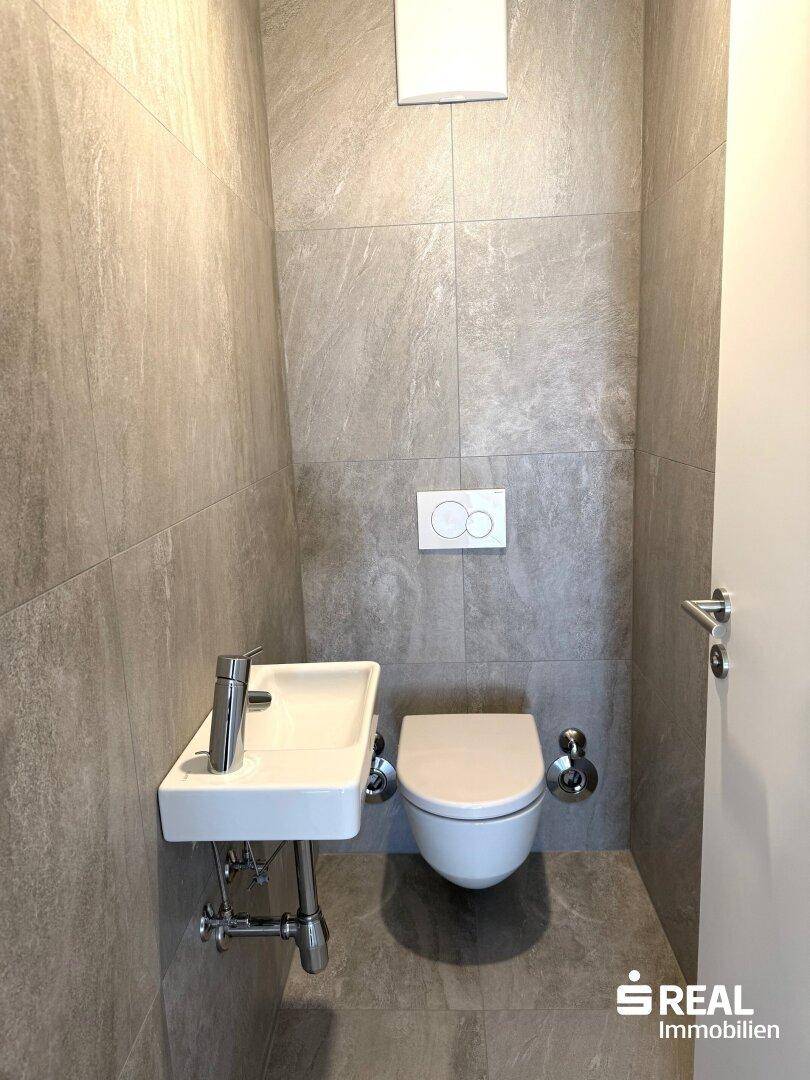 Separates Gäste-WC mit modernen grauen Fliesen, wandhängender Toilette und kleinem Handwaschbecken.