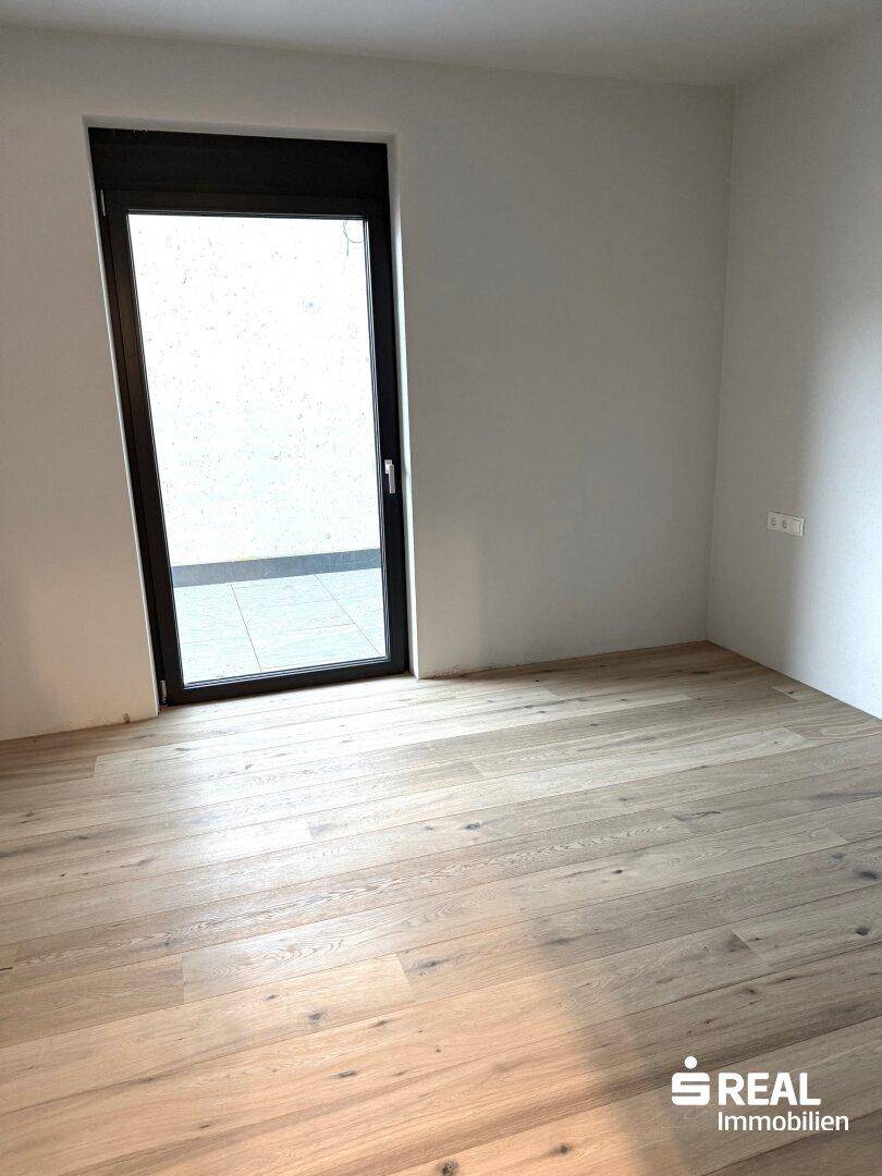 Helles Schlafzimmer mit Holzparkettboden und bodentiefem Fenster, das viel Tageslicht hereinlässt.