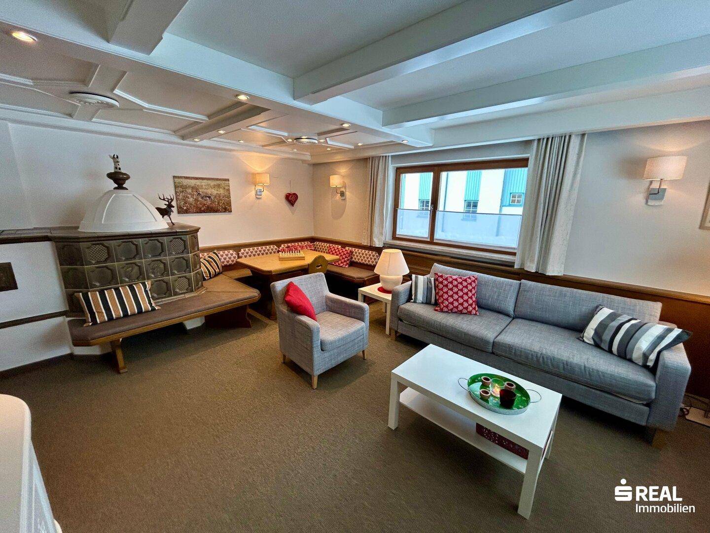 Heller Wohn- und Essbereich mit bequemen Sofas, Kachelofen und Fenstern mit Blick nach draußen.