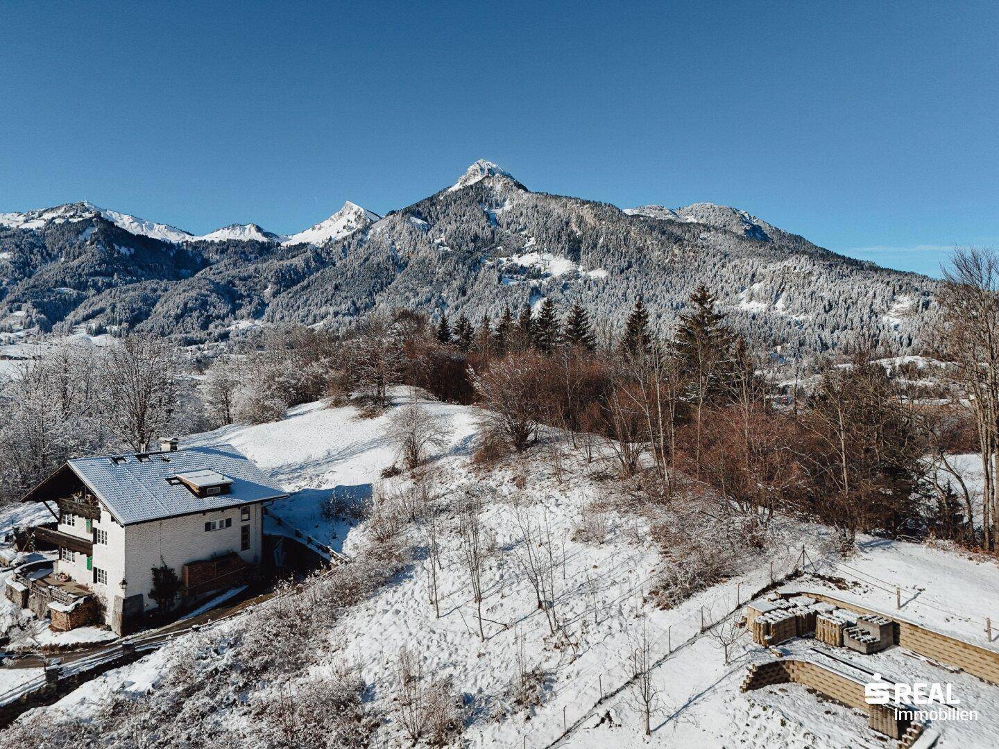 Panoramablick auf ein Haus in verschneiter Landschaft mit majestätischen Bergen im Hintergrund unter blauem Himmel.
