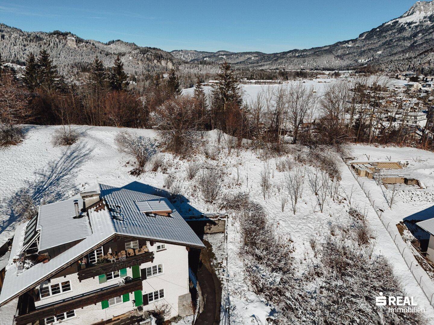 Luftaufnahme eines Hauses in verschneiter Umgebung mit Blick auf einen zugefrorenen See und Berge.