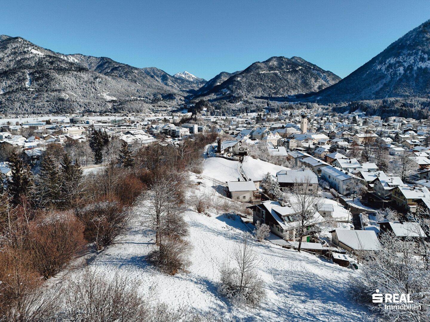 Luftaufnahme einer verschneiten Stadt in einem Tal, umgeben von schneebedeckten Bergen unter klarem Himmel.