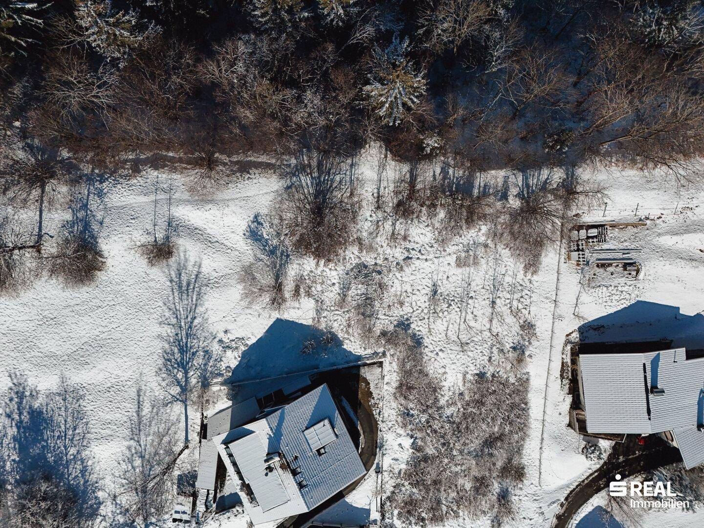 Luftaufnahme eines schneebedeckten Grundstücks, umgeben von Wald und weiteren Gebäuden in winterlicher Landschaft.