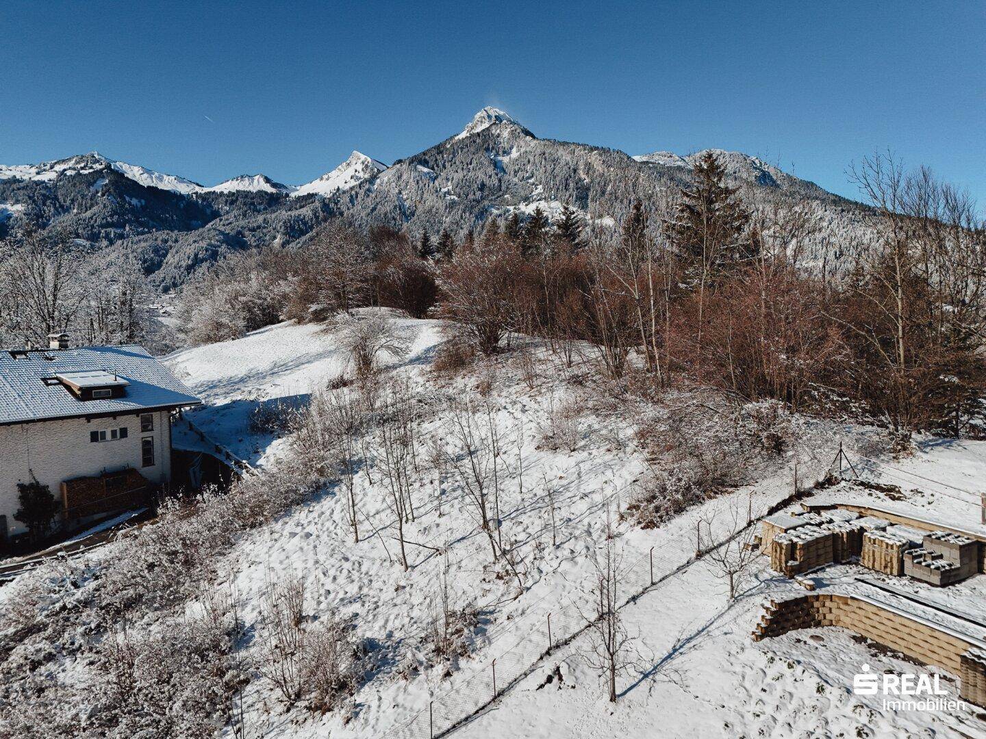 Panoramablick auf ein Haus in verschneiter Landschaft mit majestätischen Bergen im Hintergrund unter blauem Himmel.