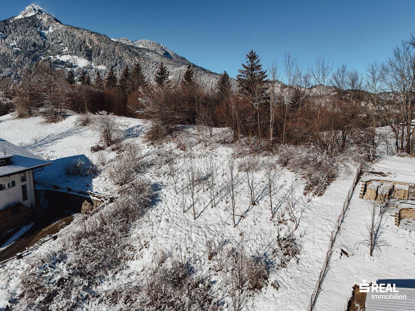 Luftaufnahme eines verschneiten Hangs mit Baustelle, teilweise sichtbarem Haus und Bergen im Hintergrund.