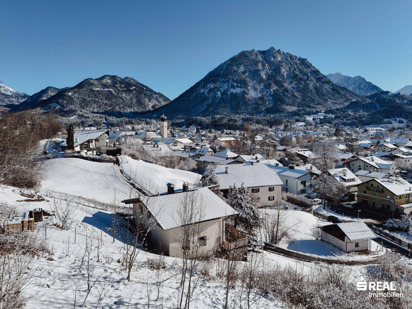 Luftaufnahme einer verschneiten Wohnsiedlung mit traditionellen Häusern und majestätischen Bergen im Hintergrund.