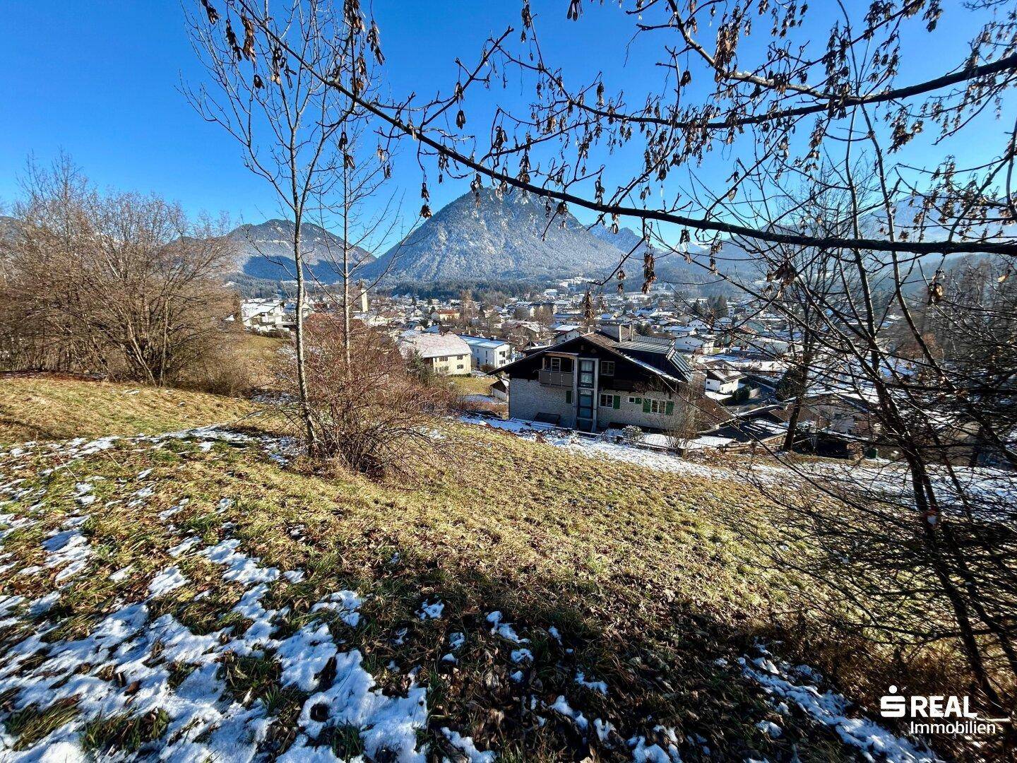 Blick über einen teilweise schneebedeckten Hang auf ein Haus, eine Stadt und die umliegenden Berge.