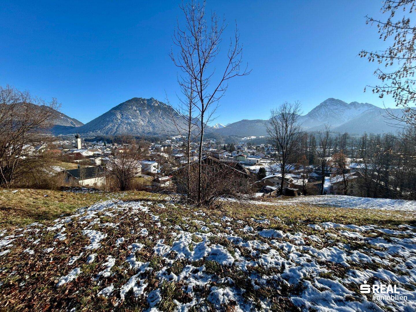 Weitblick über einen schneebedeckten Hügel auf eine malerische Stadt und die imposante Berglandschaft.