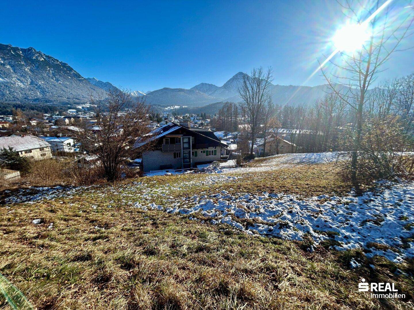 Sonniger Blick über einen teilweise schneebedeckten Hang auf ein Haus, eine Stadt und die umliegenden Berge.