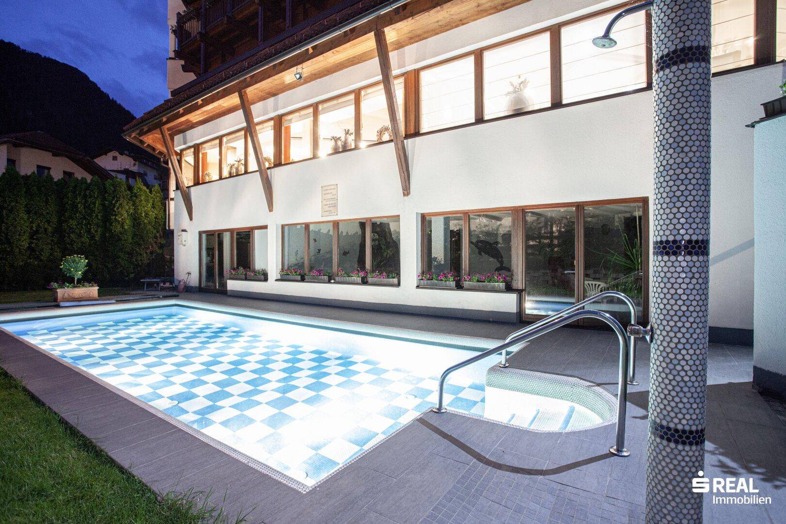 Außenpoolbereich bei Nacht mit beleuchtetem Becken und Außendusche am Gebäude.