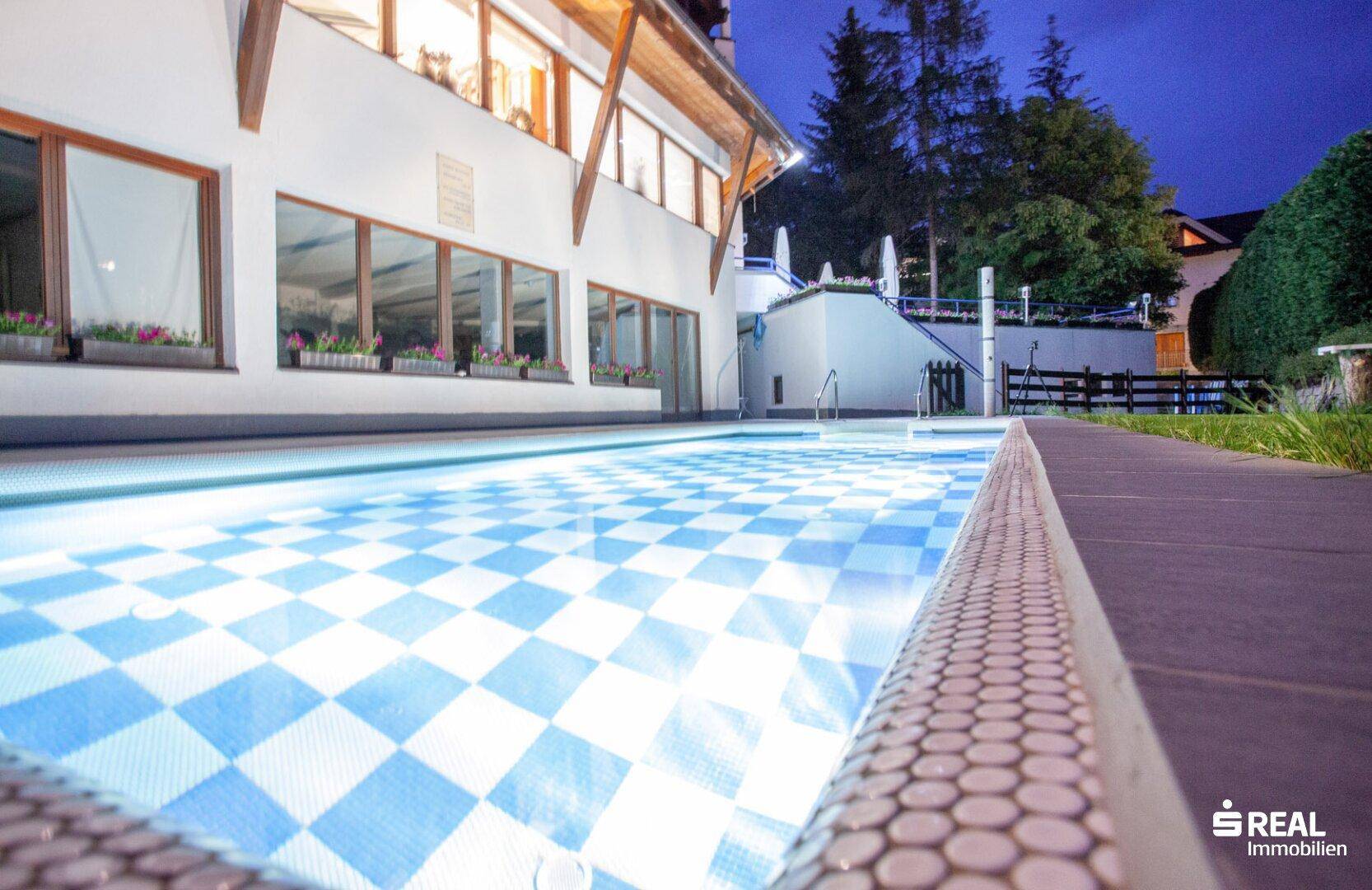 Nahaufnahme des beleuchteten Außenpools bei Nacht mit blau-weißem Fliesenmuster.