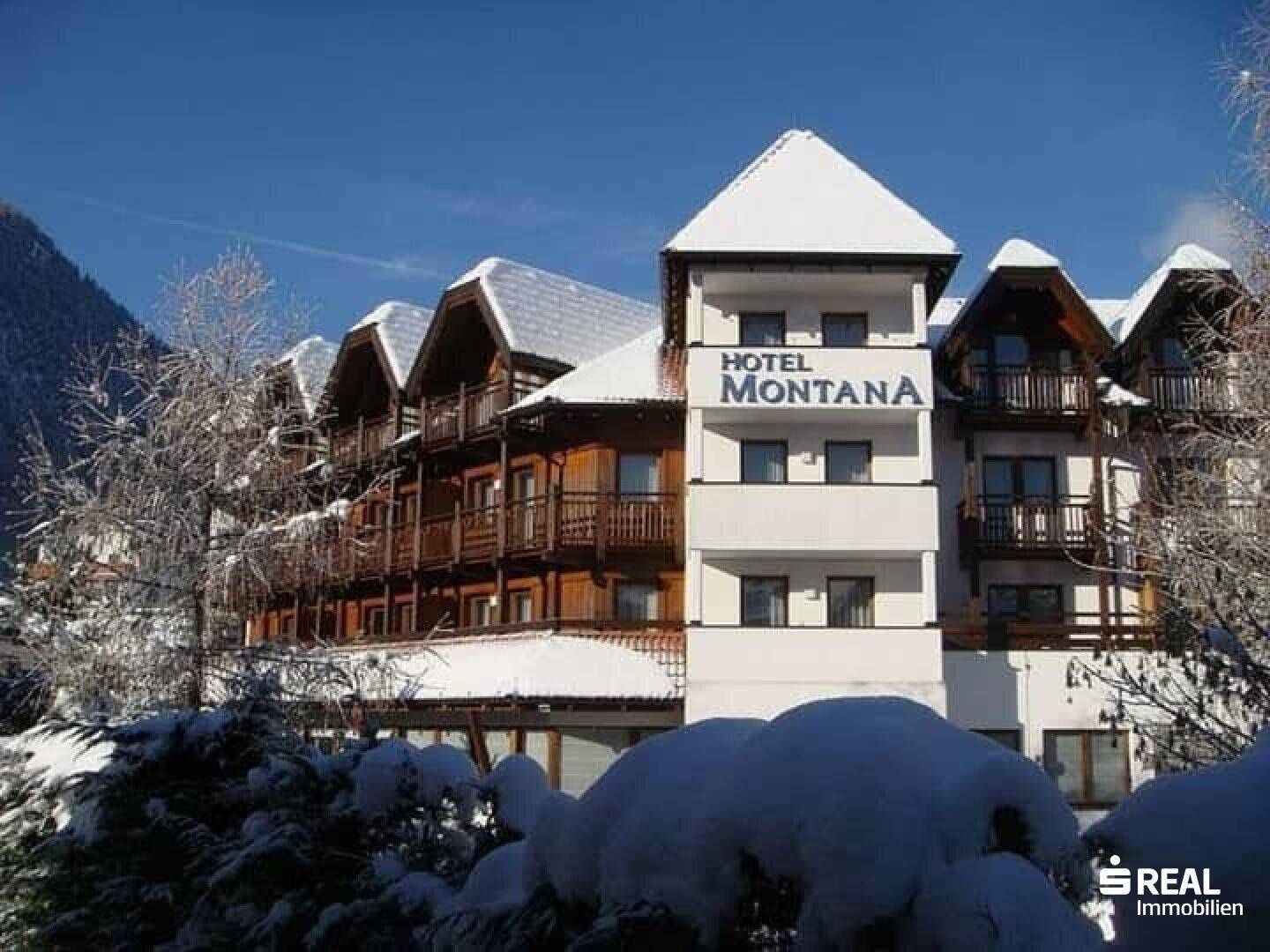 Hotelgebäude im Winter mit schneebedeckten Dächern und Balkonen vor einer Bergkulisse.