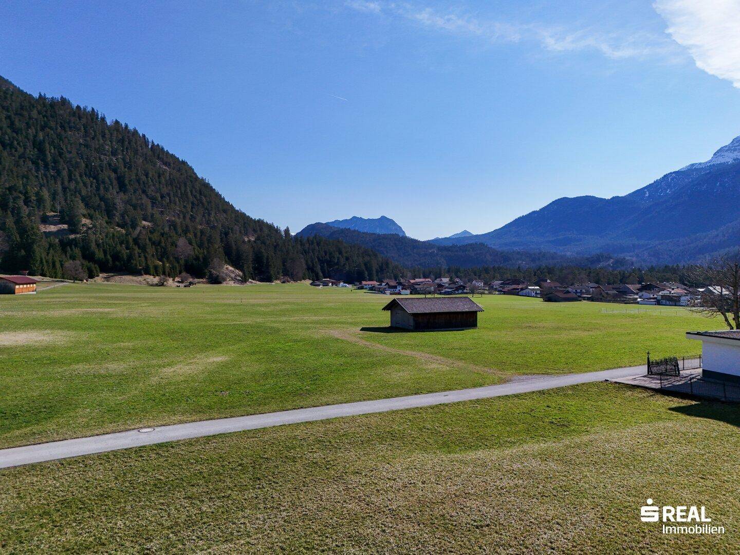 Weitläufige grüne Wiesenlandschaft mit einem kleinen Holzschuppen und majestätischen Bergen im Hintergrund unter blauem Himmel.