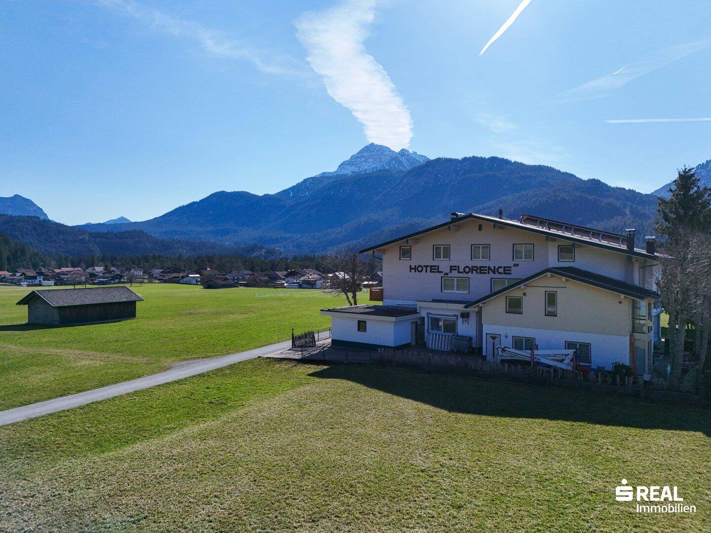 Hotelgebäude "Florence" inmitten einer grünen Wiese, umgeben von schneebedeckten Bergen unter strahlend blauem Himmel.