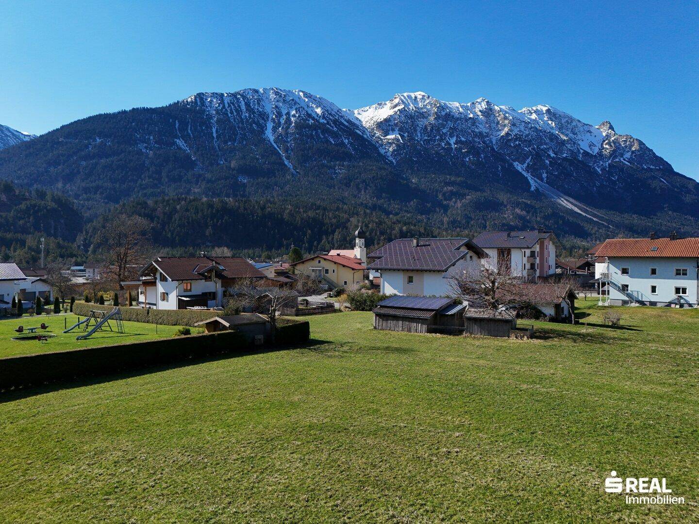 Malerisches Dorf mit traditionellen Häusern und Kirchturm, eingebettet in eine grüne Landschaft vor schneebedeckten Bergen.