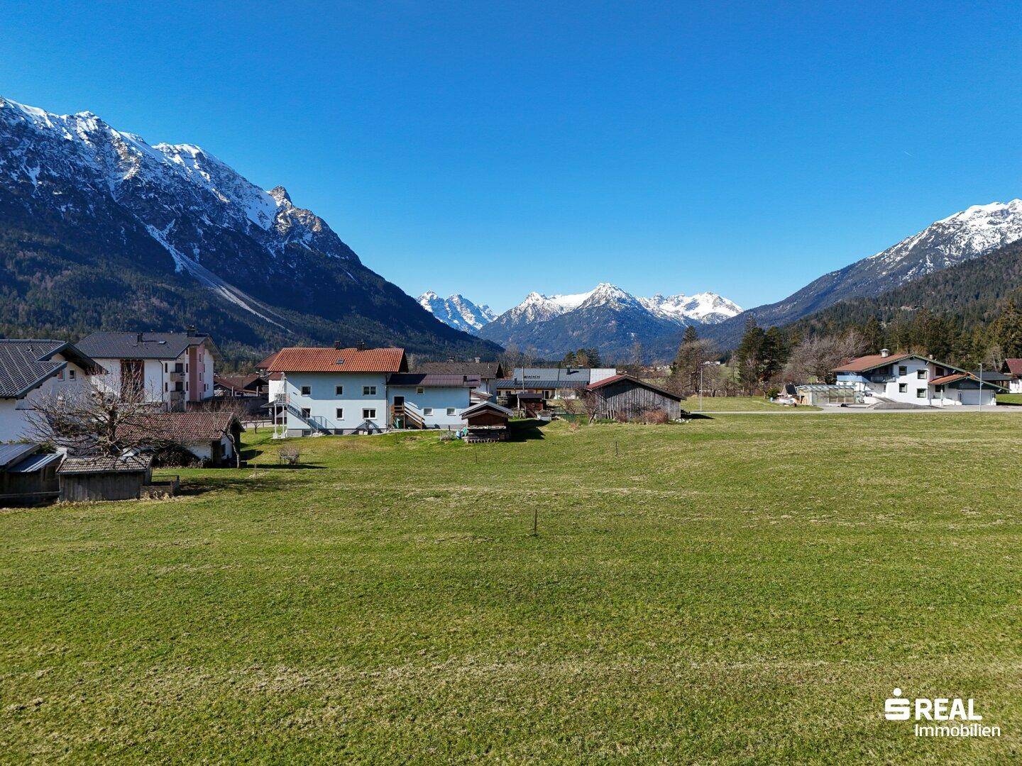 Weitläufiger Blick über ein grünes Tal mit verstreuten Häusern und beeindruckenden schneebedeckten Berggipfeln am Horizont.