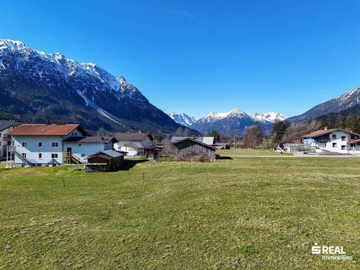 Weitläufiger Blick über ein grünes Tal mit verstreuten Häusern und beeindruckenden schneebedeckten Berggipfeln am Horizont.