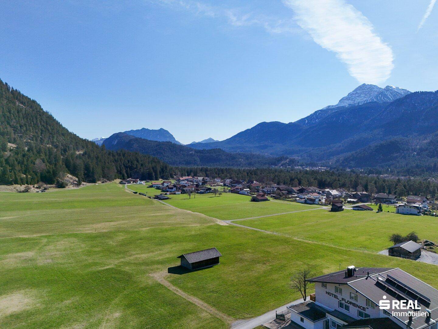Luftaufnahme des Hotel Florence und eines Schuppens in einer grünen Wiese mit Dorf und Bergen im Hintergrund.