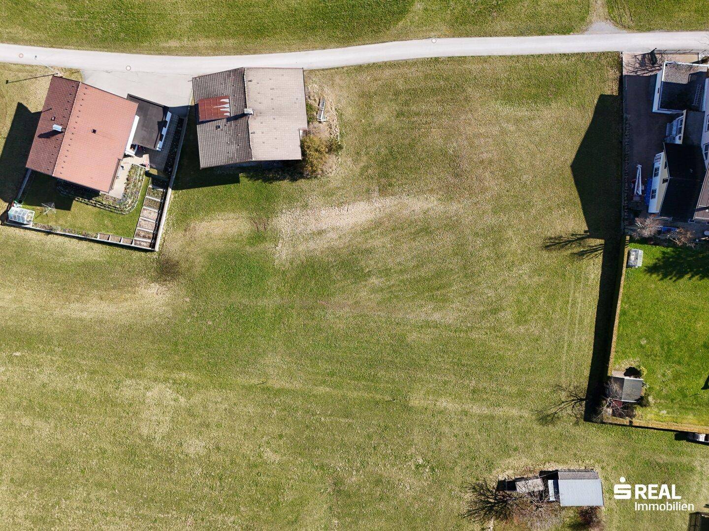 Luftaufnahme einer weitläufigen grünen Wiese, umgeben von einzelnen Häusern und Wegen in einer ruhigen Landschaft.