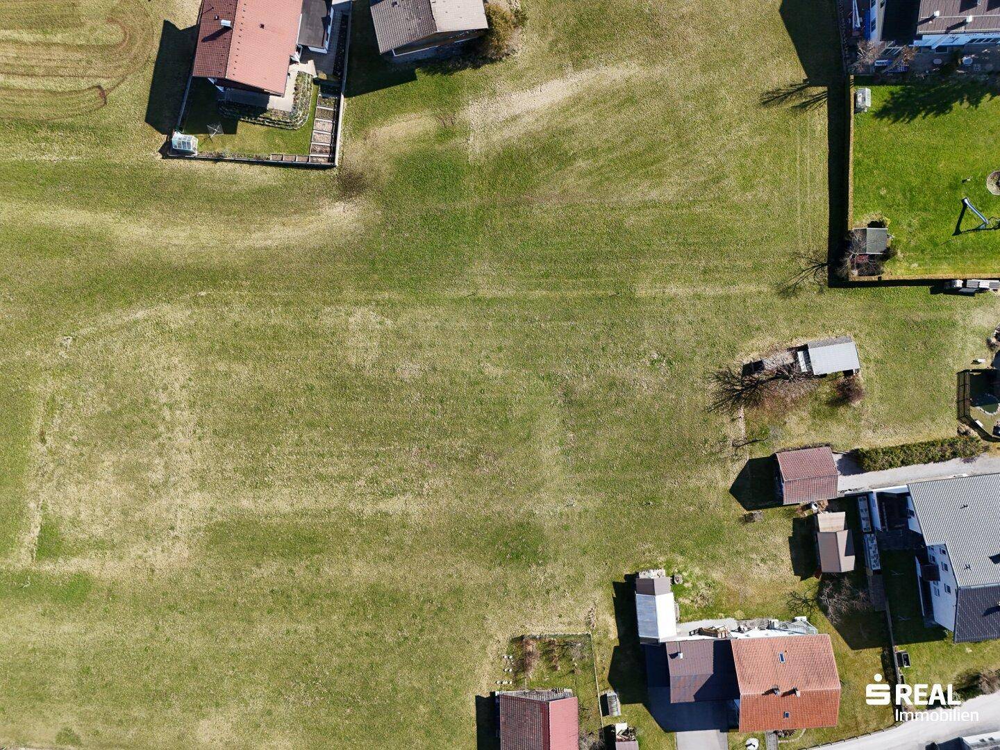 Luftaufnahme einer weitläufigen grünen Wiese, umgeben von einzelnen Häusern und Wegen in einer ruhigen Landschaft.