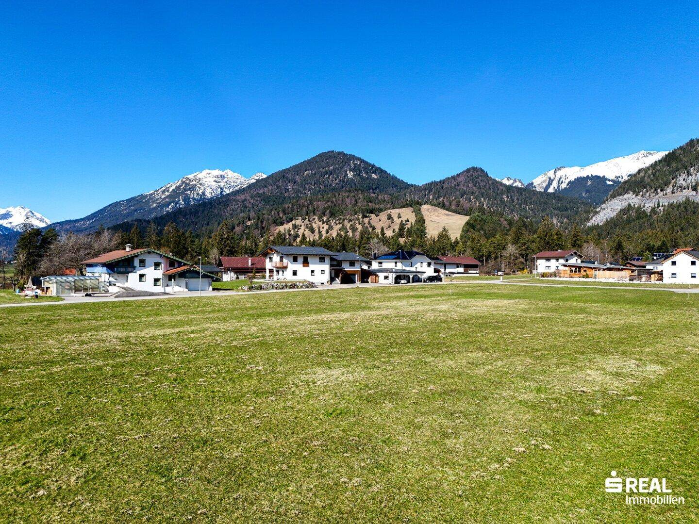Idyllische Landschaft mit schneebedeckten Bergen, grünen Feldern und vereinzelten Häusern.