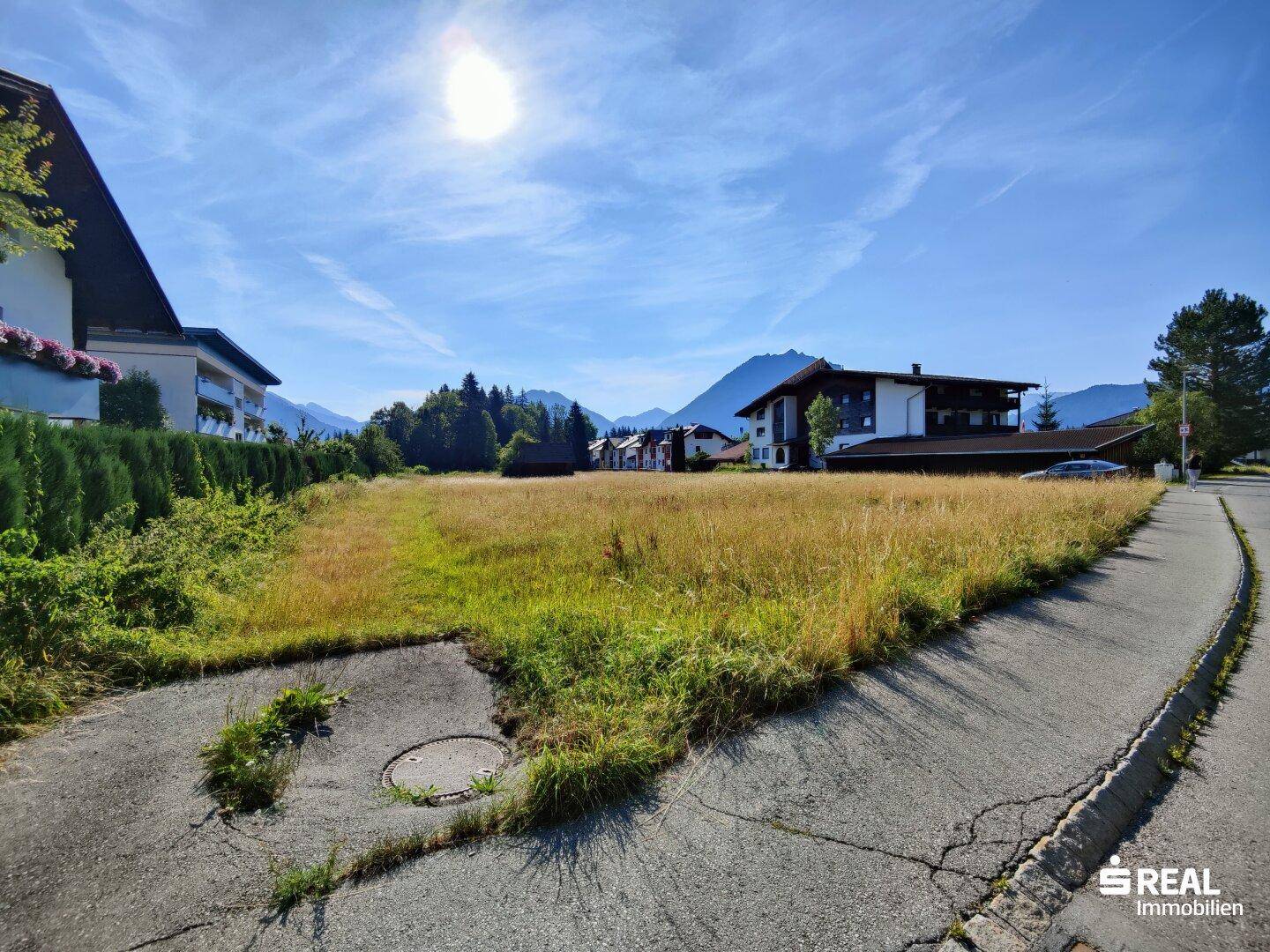Grundstück mit Wiese und angrenzender Straße, umgeben von Häusern und Bergen im Sonnenschein.